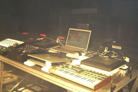 Een donker podium met dj-materiaal: mengpaneel, laptops en een keyboard; op het laptopscherm het kruislogo van Justice